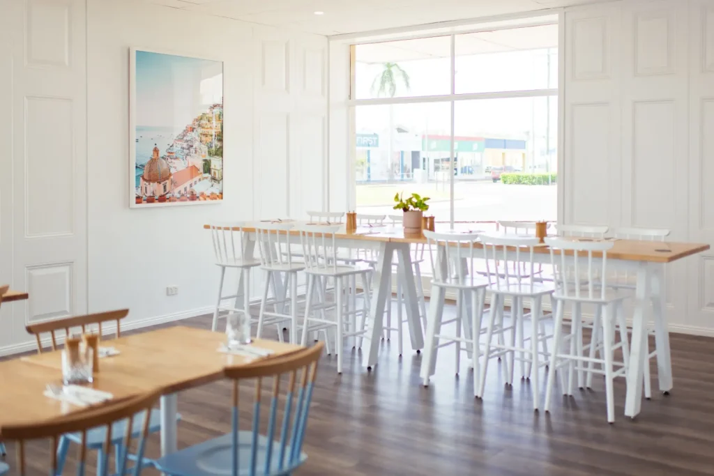 Bright and modern Italian restaurant interior with wooden tables, white chairs, large windows, and coastal artwork on the wall